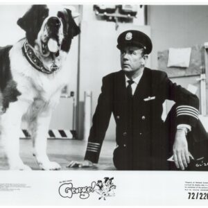 Large fluffy dog with tongue out sits beside a man in a naval captain’s uniform kneeling on the floor in a studio setting.