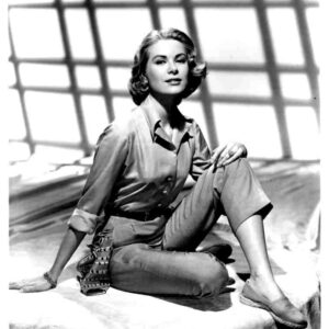 Vintage black-and-white portrait of a woman seated on a surface, wearing a buttoned blouse and cropped pants, with geometric window shadows behind her.