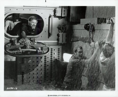 Black-and-white behind-the-scenes photo: a man leans through a round hatch and grips a control wheel, while two others brace against a wall as water sprays over them on a film set (Universal City Studios).