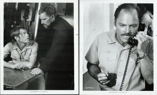 Two men in work uniforms converse in a control-room setting; one sits at a table, the other leans in from the side.