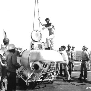 Crew of sailors and engineers work on a small submersible on a ship deck, with a man on top guiding a hoist.