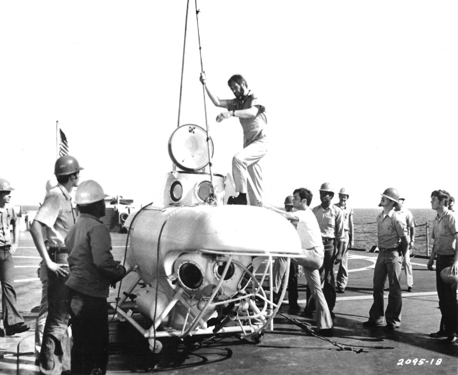 Crew of sailors and engineers work on a small submersible on a ship deck, with a man on top guiding a hoist.