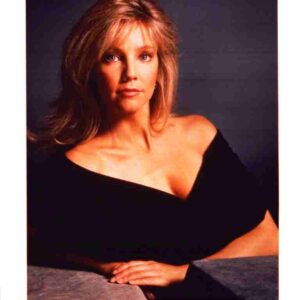 Studio portrait of a blonde woman in a black off-the-shoulder top, seated with hands folded on a gray surface.