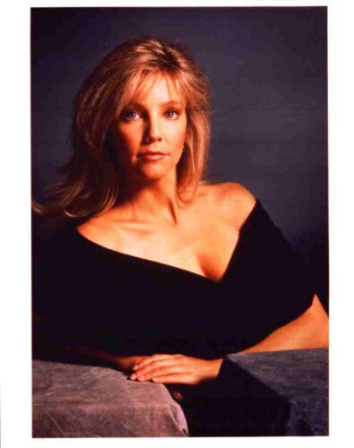 Studio portrait of a blonde woman in a black off-the-shoulder top, seated with hands folded on a gray surface.