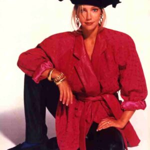 Woman in a red belted blazer with rolled sleeves, black wide-brimmed hat, and chunky jewelry posing with one knee up in a studio setting.