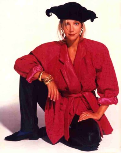 Woman in a red belted blazer with rolled sleeves, black wide-brimmed hat, and chunky jewelry posing with one knee up in a studio setting.