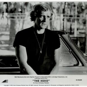 Black-and-white promotional still: man in a dark T-shirt with a pendant, leaning beside a car outdoors.