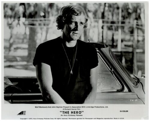 Black-and-white promotional still: man in a dark T-shirt with a pendant, leaning beside a car outdoors.