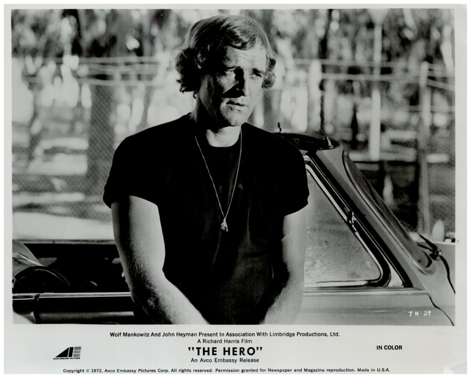 Black-and-white promotional still: man in a dark T-shirt with a pendant, leaning beside a car outdoors.