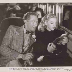 Vintage movie still of a man in a checked suit and a blonde-haired woman reading a magazine inside a train car.