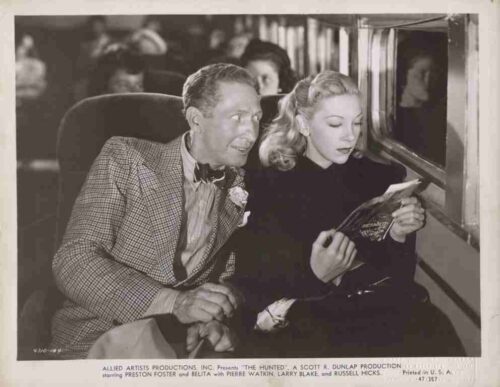 Vintage movie still of a man in a checked suit and a blonde-haired woman reading a magazine inside a train car.