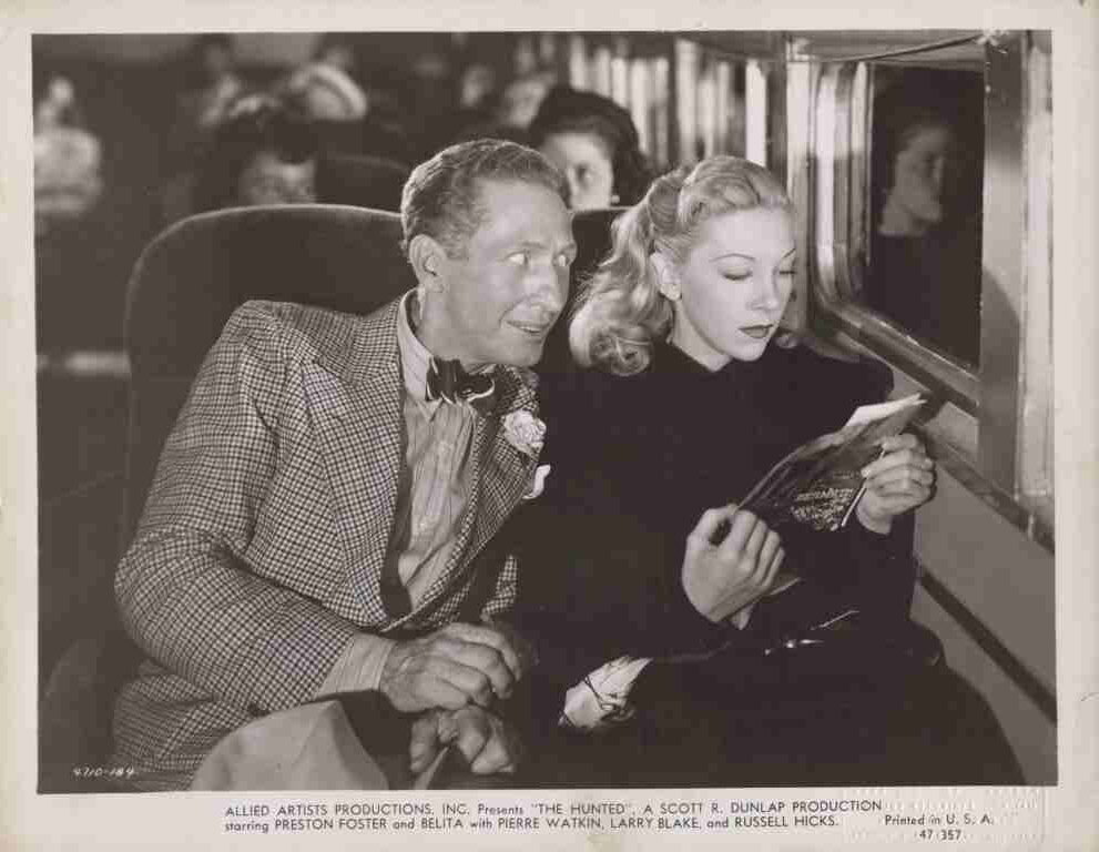 Vintage movie still of a man in a checked suit and a blonde-haired woman reading a magazine inside a train car.