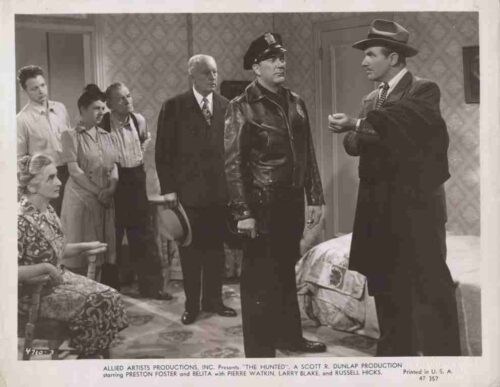 Black-and-white film still from The Hunted: a sheriff in a leather jacket confronts a man in a hat while others observe in a patterned living room.