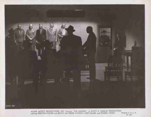 Black-and-white film still showing a lineup of men in suits on a stage, with guards and a silhouette-filled audience in the foreground.