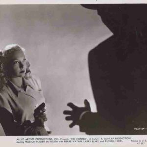 A woman in a trench coat holds a pistol, glancing toward a large dark silhouette with an reaching hand in the foreground; tense studio scene.