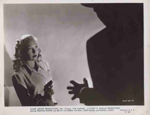 A woman in a trench coat holds a pistol, glancing toward a large dark silhouette with an reaching hand in the foreground; tense studio scene.