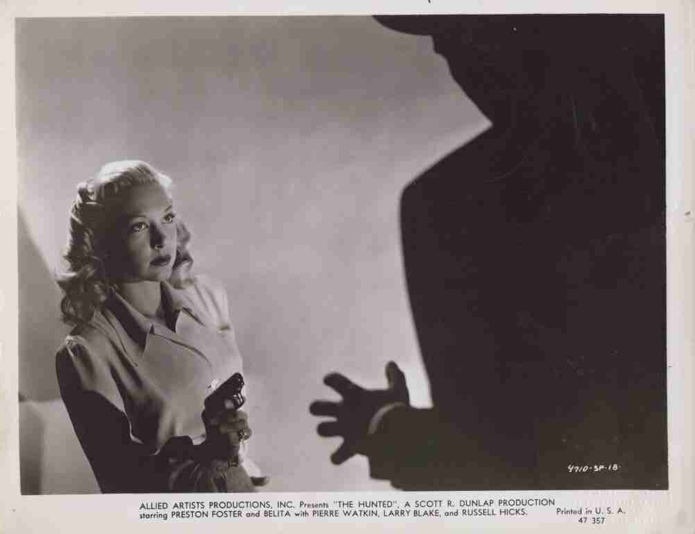 A woman in a trench coat holds a pistol, glancing toward a large dark silhouette with an reaching hand in the foreground; tense studio scene.