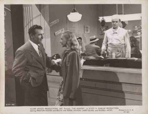 Promotional still from The Hunter: a man in a suit talks with a woman in a coat beside a diner counter; a waiter stands behind the counter.