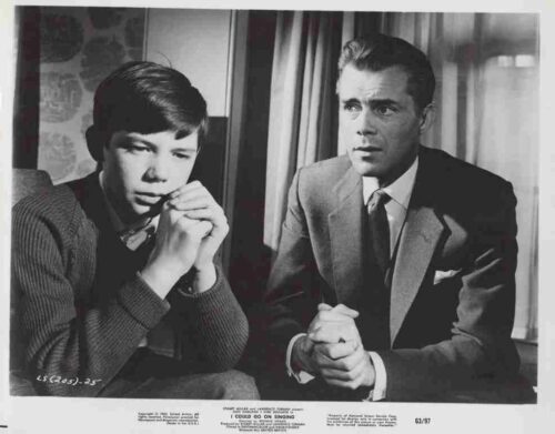 Two men sit side by side in a black‑and‑white photo, one resting his chin on his hands and the other with hands clasped, wearing a sweater and a suit.