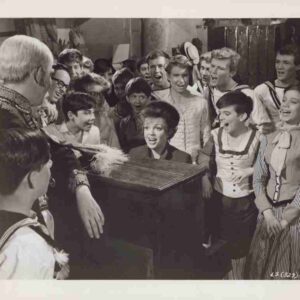 A young woman at a piano singing or performing while a crowd of smiling children in vintage outfits surrounds her.