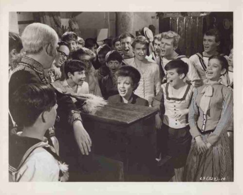 A young woman at a piano singing or performing while a crowd of smiling children in vintage outfits surrounds her.