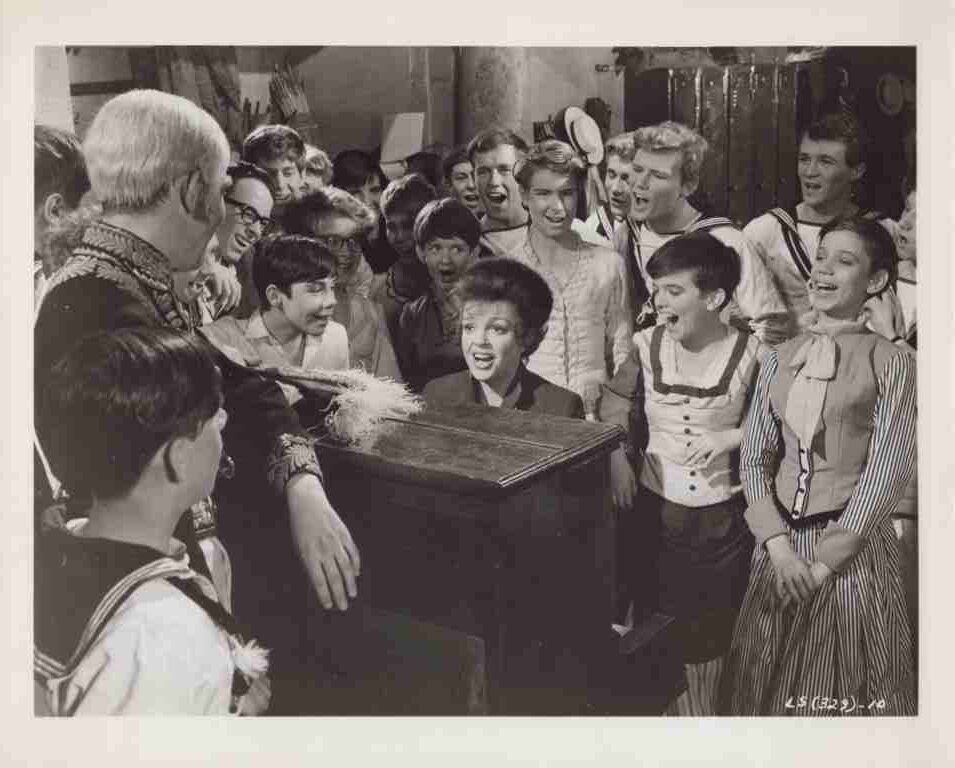 A young woman at a piano singing or performing while a crowd of smiling children in vintage outfits surrounds her.