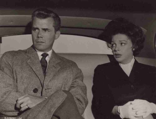 Man in a tweed suit and a woman in a dark coat sit in the backseat of a car, looking off to the left.