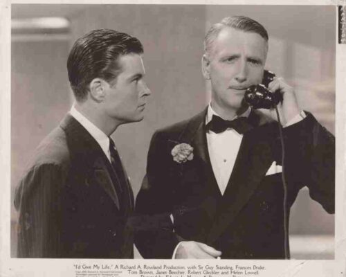 Black-and-white movie still of two men in tuxedos, one on a telephone, in a formal scene.