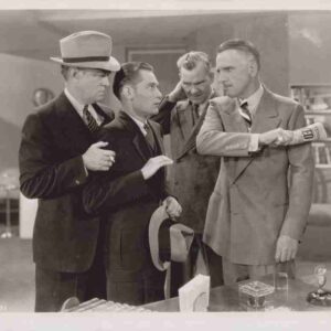 Four men in 1940s-style suits stand in an office; one man extends his arm with a 'FED' armband while three others observe, one holding a hat.