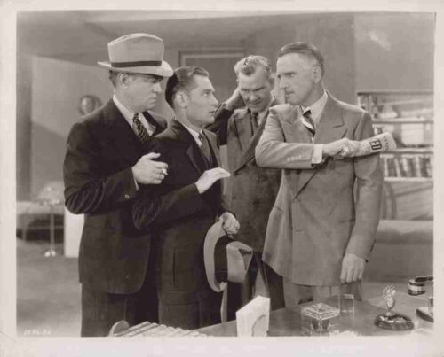 Four men in 1940s-style suits stand in an office; one man extends his arm with a 'FED' armband while three others observe, one holding a hat.