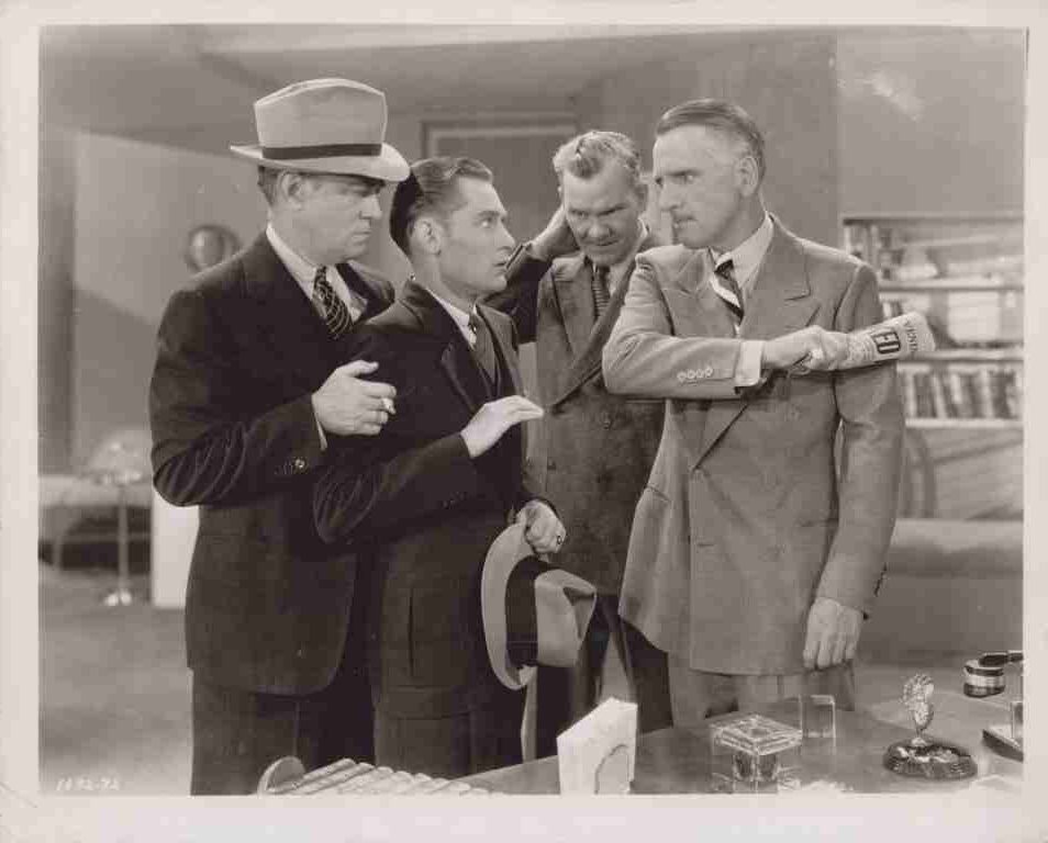 Four men in 1940s-style suits stand in an office; one man extends his arm with a 'FED' armband while three others observe, one holding a hat.