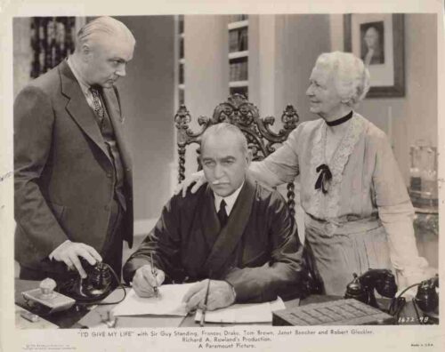 Man seated at a desk signs a document while two colleagues stand beside him in a vintage film set, ornate chair behind him and old-style desk accessories visible.