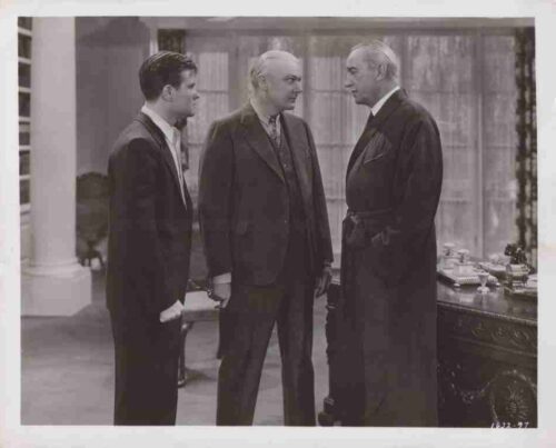 Three men in a stately room stand in a tense discussion, the middle man bald and in a suit. The man on the right wears a robe or gown.