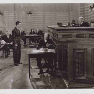 In a courtroom, a judge sits at the elevated bench while a man signs documents at a table in front of him, with people standing and seated in the background.