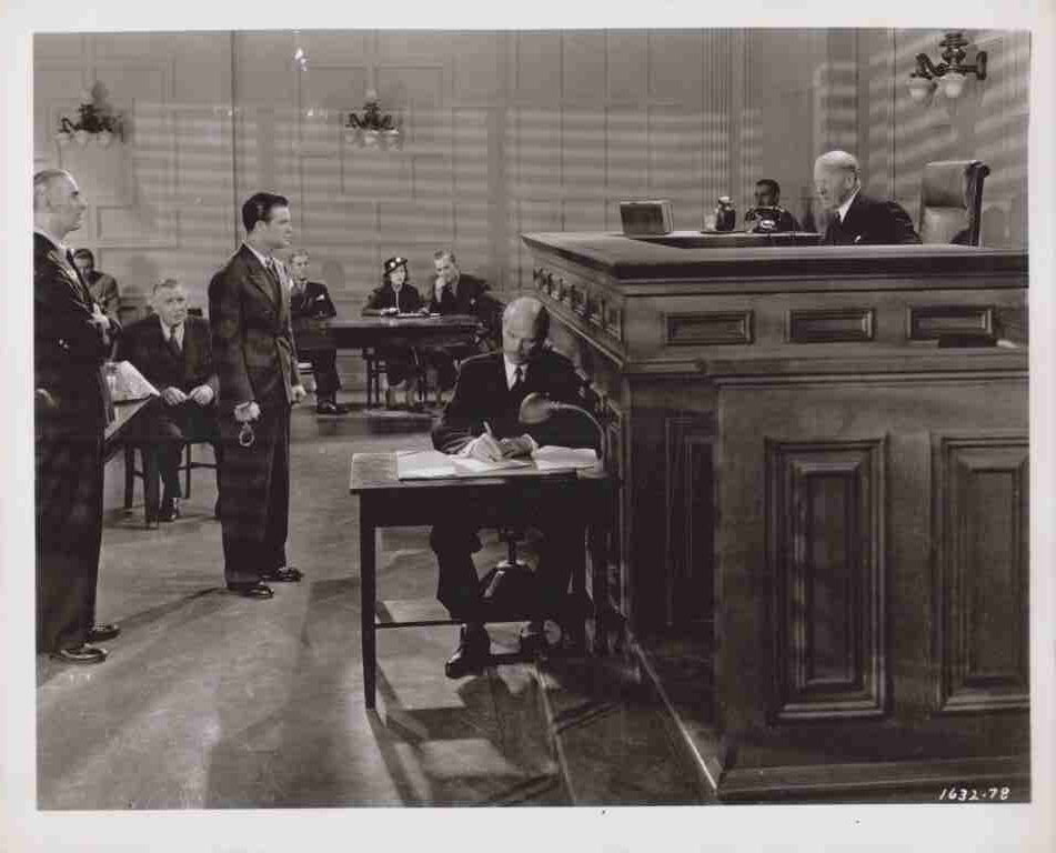 In a courtroom, a judge sits at the elevated bench while a man signs documents at a table in front of him, with people standing and seated in the background.