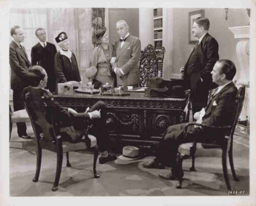 A formal office scene with two men seated at an ornate desk facing a group of standing colleagues.