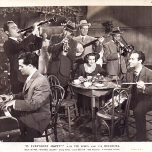 Black-and-white photo of Ted Lewis and his orchestra in a dining room: pianist on the left, drummer on the right, four horn players standing behind a table where a woman in a hat sits among cups and dessert; caption reads 'Is Everybody Happy?'