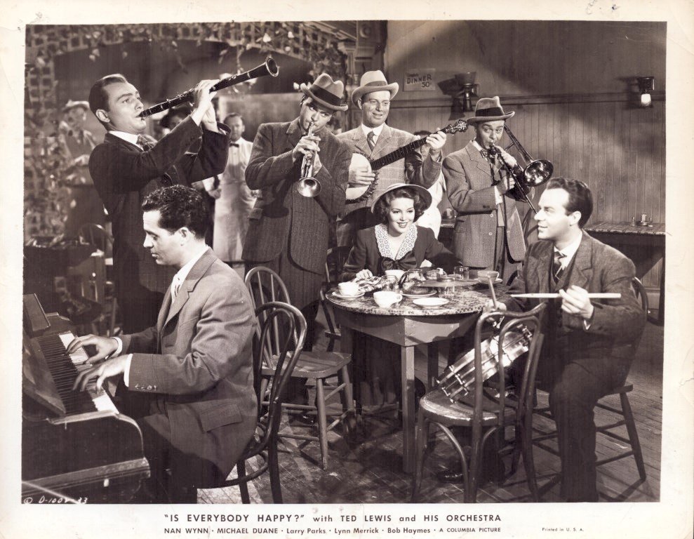 Black-and-white photo of Ted Lewis and his orchestra in a dining room: pianist on the left, drummer on the right, four horn players standing behind a table where a woman in a hat sits among cups and dessert; caption reads 'Is Everybody Happy?'