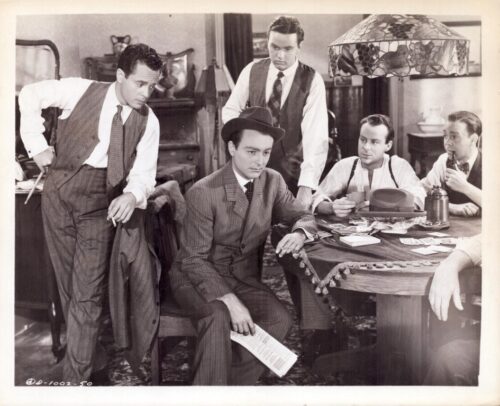 Five men in 1930s-style suits gather around a round card table, watching a man in a hat who holds cards. A lamp hangs overhead and a cluttered parlor surrounds them.