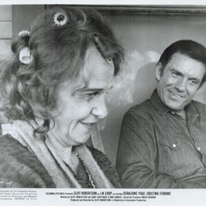 Black-and-white photo of a smiling woman with curlers in her hair and a man in a denim shirt sitting on a porch, smiling at each other.