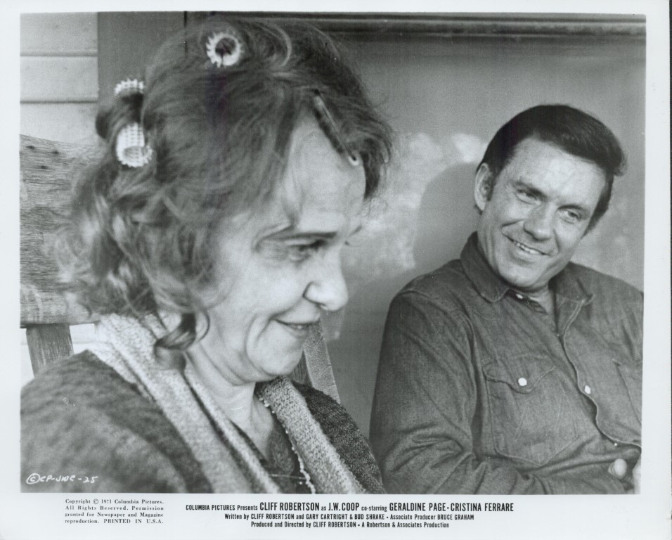 Black-and-white photo of a smiling woman with curlers in her hair and a man in a denim shirt sitting on a porch, smiling at each other.