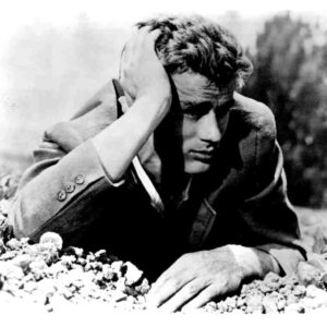 Black-and-white photo of a man in a suit resting with his head in one hand, lying among rocks outdoors, looking somber.