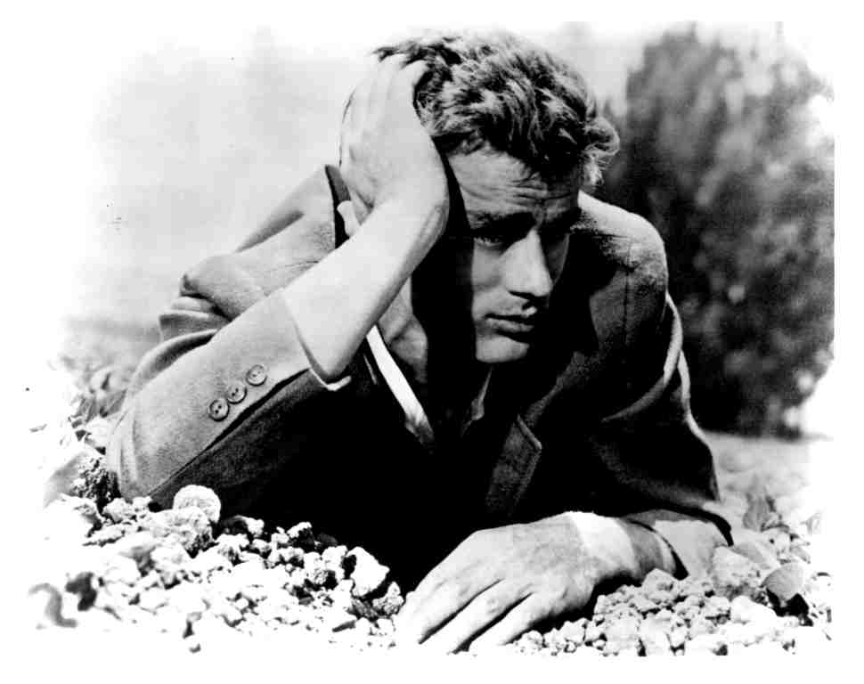 Black-and-white photo of a man in a suit resting with his head in one hand, lying among rocks outdoors, looking somber.