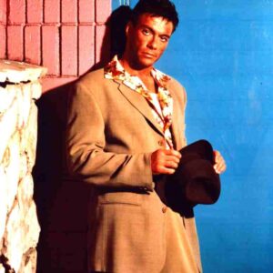 Man in a beige suit leaning against a pink and blue wall, holding a dark hat.