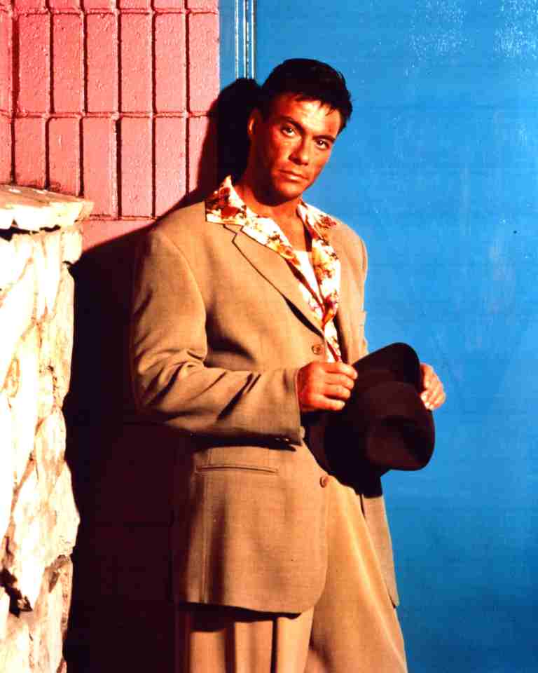 Man in a beige suit leaning against a pink and blue wall, holding a dark hat.