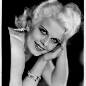 Black-and-white portrait of a smiling woman with short blonde hair, resting her head on folded hands, wearing earrings and a bracelet.