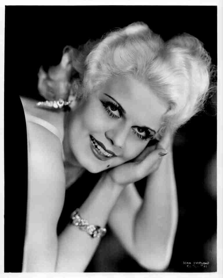 Black-and-white portrait of a smiling woman with short blonde hair, resting her head on folded hands, wearing earrings and a bracelet.