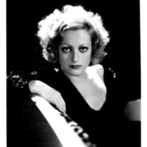 Black-and-white portrait of a woman with short, curly blonde hair wearing a dark V-neck dress, gazing at the camera.