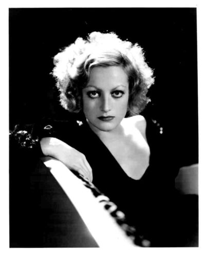 Black-and-white portrait of a woman with short, curly blonde hair wearing a dark V-neck dress, gazing at the camera.