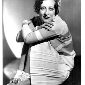Black-and-white portrait of a woman with short wavy hair, wearing a striped dress, sitting with arms around her knees and smiling.